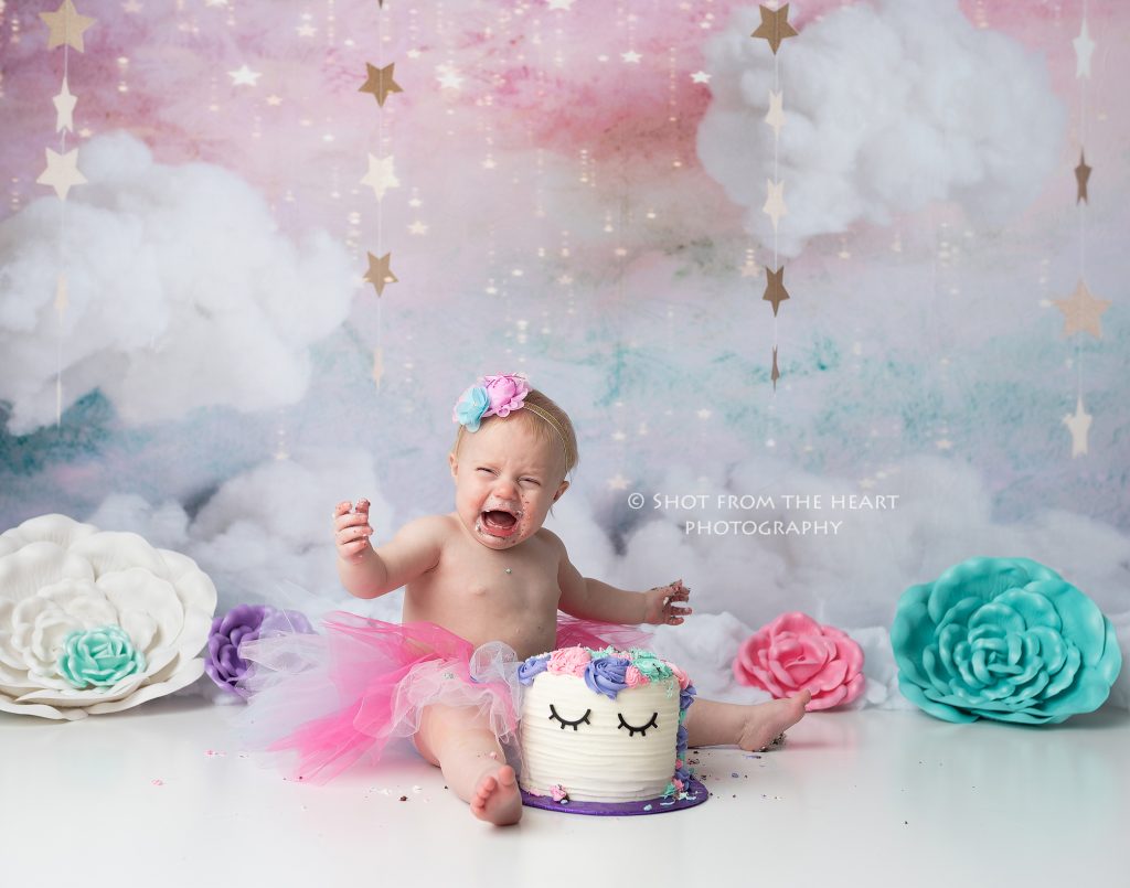 Unicorn birthday cake smash photography session with pink, purple, teal, and white colors. Crying baby with birthday cake. 