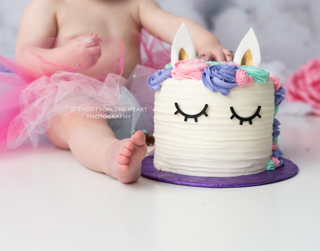 Unicorn birthday cake smash photography session with pink, purple, teal, and white colors.  Close up of toes and birthday cake. 