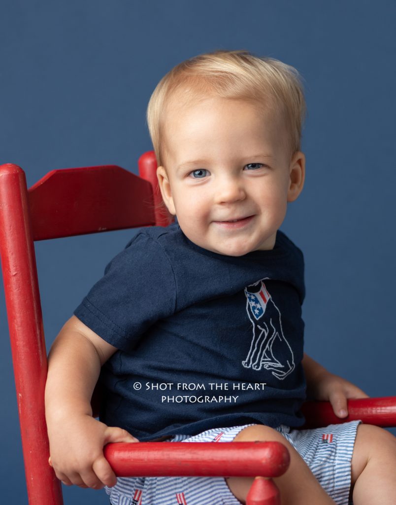 first birthday portraits. baby in red rocking chair on navy blue backdrop.